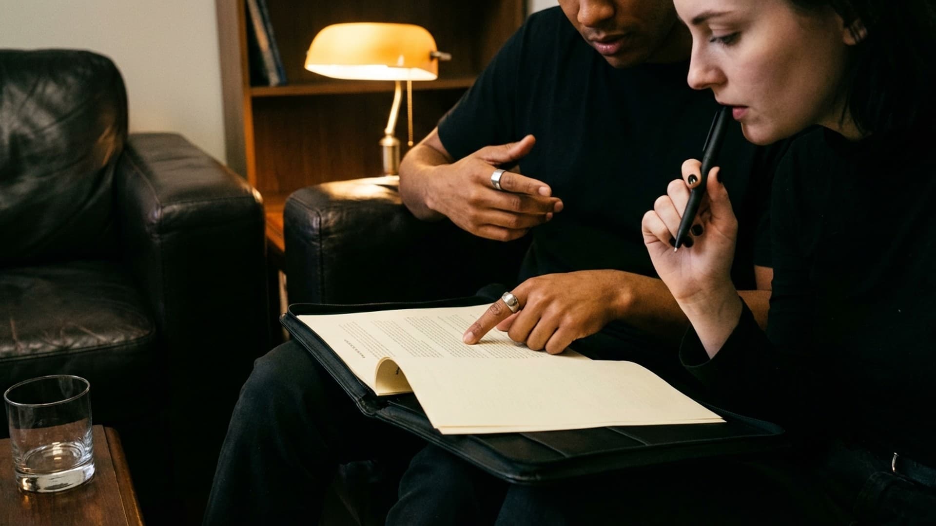 Two people sitting close, one writing in a notebook while the other watches, lit by warm amber light.